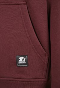 Felpa bordeaux in tessuto morbido con tasca a marsupio frontale e una toppa quadrata nera con scritte bianche e una stella.