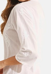 Witte, lichtgewicht blouse met driekwart mouwen. Heeft bloemenborduursels op de mouwen en aan de voorkant. Zachte, gestructureerde stof.