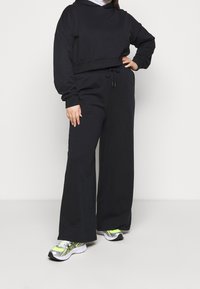 Svart kort hoodie med långa ärmar och dragsko, kombinerad med vida svarta joggers. Modellen bär silvriga och neongröna sneakers.