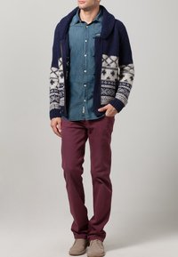 Marinblå stickad cardigan med vita och blå mönster, lagd över en denimskjorta, tillsammans med vinröda byxor och beige skor.