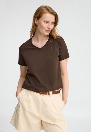 Donna con maglietta marrone a scollo a V e pantaloncini beige a vita alta con tasche, in piedi con le mani nelle tasche su uno sfondo semplice.