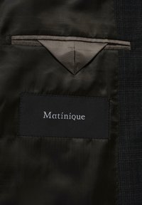 Sort satinjakkeforing med en lille indvendig lomme og et sort rektangulært mærke syet nedenfor med "Matinique" i hvid tekst.