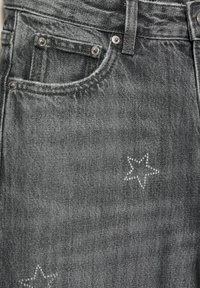 Gros plan sur la poche avant d'un jean en denim noir avec des rivets argentés et deux décorations en strass en forme d'étoile sur le tissu.