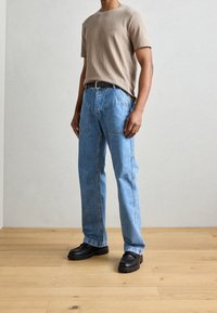 Uomo che indossa una camicia beige a maniche corte, jeans azzurri con tasche applicate, cintura nera e mocassini neri spessi, in piedi su un pavimento di legno.