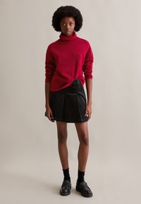 Roter Rollkragenpullover mit hochgerollten Ärmeln, am Taillenbereich geknotet; schwarzer, plissierter Rock; schwarze Slipper; vor einem neutralen Hintergrund.