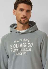 Grauer Kapuzenpullover mit weißem Text: "QUALITY GOODS S.OLIVER CO. ELEVATED CASUAL SEIT 1969." Weicher Stoff, reguläre Passform.