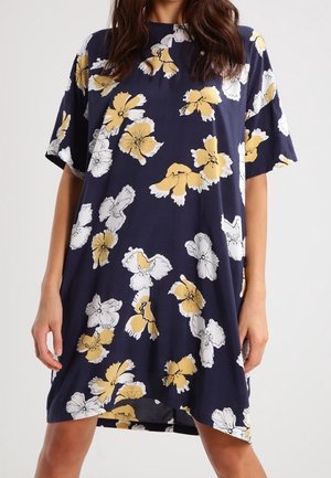 Kobieta w luźnej granatowej sukience w żółte i białe kwiaty, z krótkimi rękawami i okrągłym dekoltem.