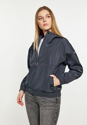 Junge Frau, die eine dunkelblaue Jacke mit Reißverschluss trägt, die Hand in der Tasche, schaut seitlich, vor einem einfarbigen hellen Hintergrund.