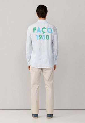 Homme debout face à un mur, portant une chemise à manches longues rayée claire avec l'inscription "FAÇO 1950" en grandes lettres dans le dos, pantalon beige et chaussures bleues.