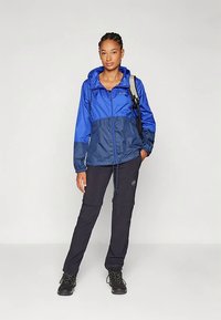 Jauna moteris vilkinti mėlyną gobtuvą turinčią striukę, juodus žygių kelnes, juodus batus ir nešanti pilką kuprinę prieš paprastą foną.