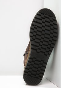 Brązowy zamszowy but z teksturowaną czarną gumową podeszwą, z bocznym zamkiem i minimalistycznym wzornictwem. Podeszwa ma głębokie rowki dla lepszej przyczepności.