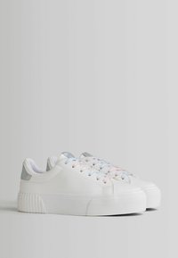 White sneakers with a smooth finish, pastel laces (pink, blue) and metallic accents, textured sole, and raised heel.