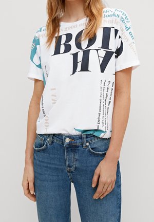 Personne portant un t-shirt blanc graphique avec un texte noir et turquoise, associé à un jean bleu, sur un fond clair uni.