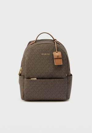 Brown patterned backpack with gold hardware, dual zipper, top handle, and front pocket. Features a branding tag and textured fabric.