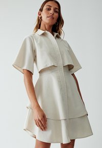 Beige, jurk met korte mouwen met een cropped bovenlaag en een uitlopende rok. Voorzien van een overhemdkraag en knoopsluiting aan de voorkant.