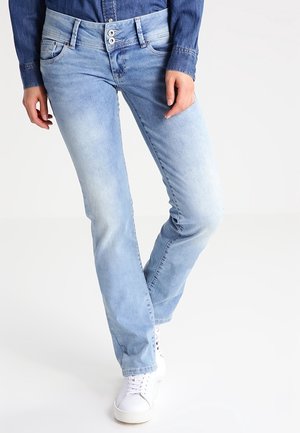 Person trägt hellblaue Jeans mit geradem Bein und Doppelknopfverschluss, dunkelblaues Denimhemd und weiße Sneakers und steht vor weißem Hintergrund.