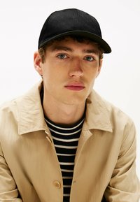 Gorra negra con patrón texturizado, combinada con una chaqueta beige y una camiseta de rayas en blanco y negro. Fondo neutro, centrado en la parte superior del cuerpo.