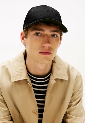 Cappello nero con motivo testurizzato, indossato con una giacca beige e una camicia a righe bianco e nero. Sfondo neutro, in primo piano la parte superiore del corpo.