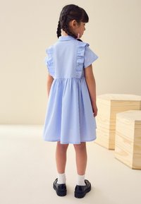 Blauwe gingham jurk met gerimpelde mouwen, getailleerde taille en uitlopende rok. Draagt witte sokken en zwarte lakleren schoenen.