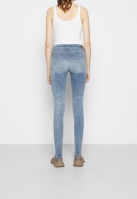 Donna con jeans skinny azzurri chiari, top bianco senza maniche e sneakers beige, in piedi contro un muro bianco semplice, vista da dietro.