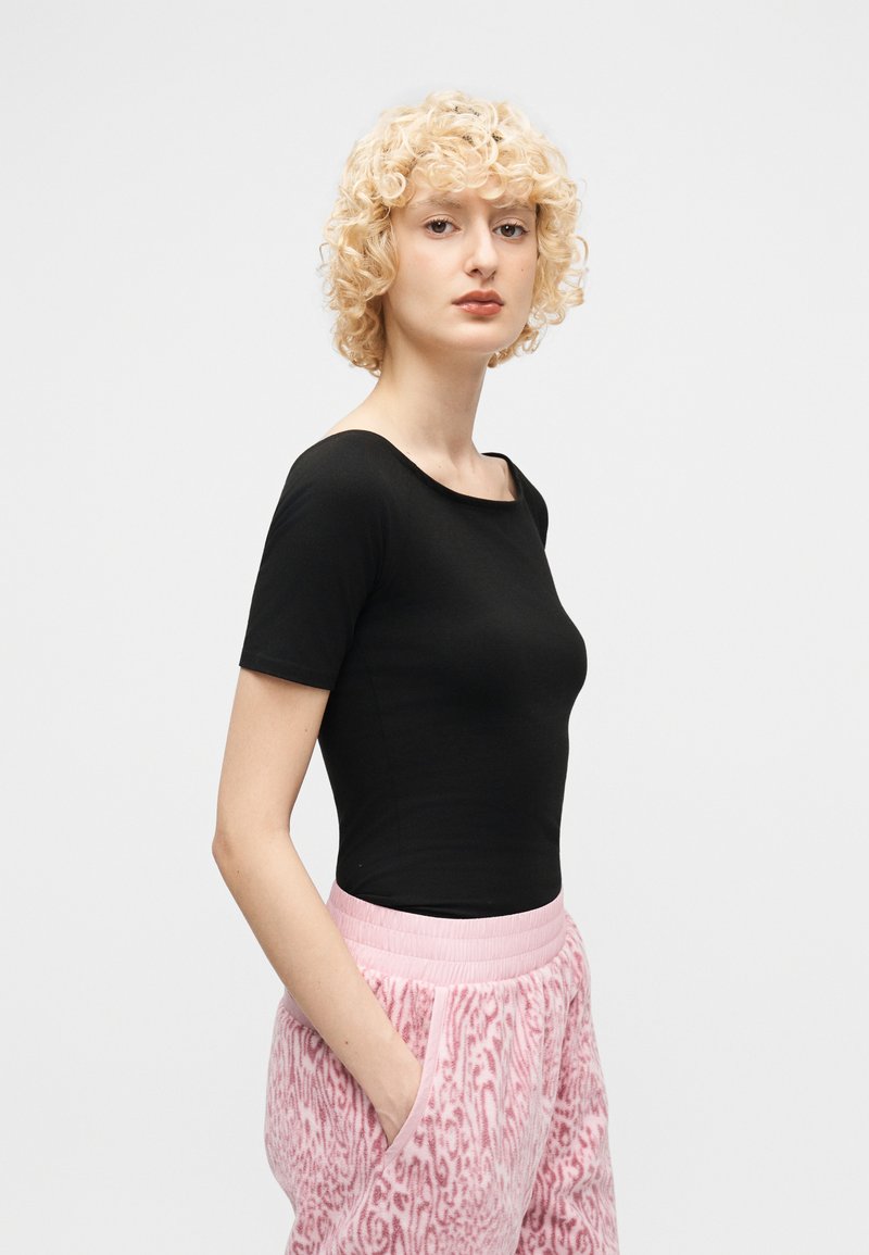Young person with short curly blonde hair wearing a fitted black short-sleeve top and pink patterned pants, standing with hands in pockets.