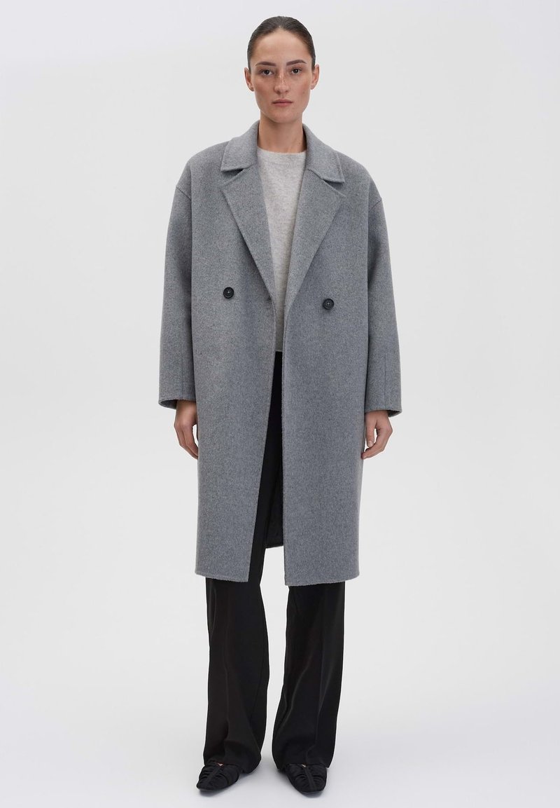 Vrouw in een lange grijze double-breasted jas over een lichtgrijze trui, met zwarte wijde broek en zwarte puntige schoenen, staande tegen een witte achtergrond.