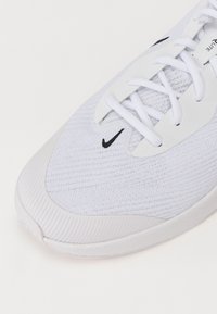 Bílé sportovní boty s síťovinovým svrškem, bílými tkaničkami, gumovou podešví a malým černým logem Nike v blízkosti špičky.