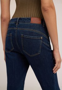 Detail osoby nosící tmavě modré džínové kalhoty a hnědé žebrované tílko bez rukávů, zobrazující zadní kapsy a prošitou koženou značkovou štítek.