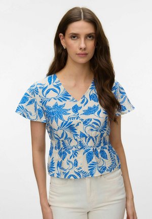 Vrouw met lang bruin haar draagt een korte mouw beige blouse met V-hals en blauwe bloemenprints, gecombineerd met witte broek, tegen een effen achtergrond.