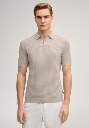 Man draagt een beige polo met korte mouwen en een getextureerde stof, gecombineerd met een witte broek, staand tegen een egale lichte achtergrond.