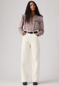 Γυναίκα με μακριά σγουρά μαλλιά που φοράει ροζ πουκάμισο με βολάν και καρό μοτίβο, λευκό παντελόνι φαρδύ με ψηλή μέση, μαύρη ζώνη και μαύρες μπότες, με τα χέρια στις τσέπες.