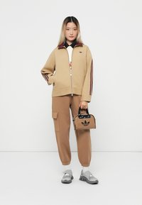 Giacca zip-up beige con strisce bordeaux, pantaloni cargo marroni e una piccola borsa nera con logo Adidas. Le sneakers grigie completano l'outfit.
