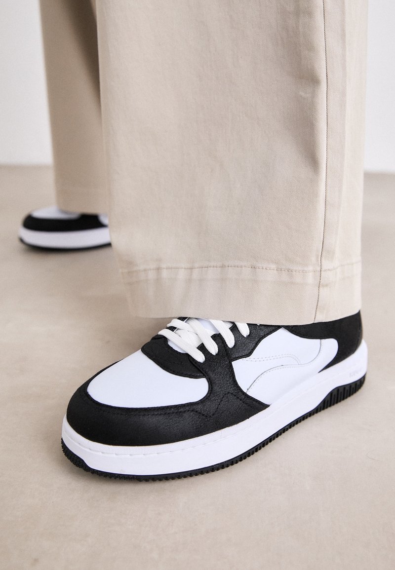 Baskets noir et blanc avec une tige en cuir texturé, une semelle robuste, des lacets blancs et un design distinctif avec des accents incurvés.