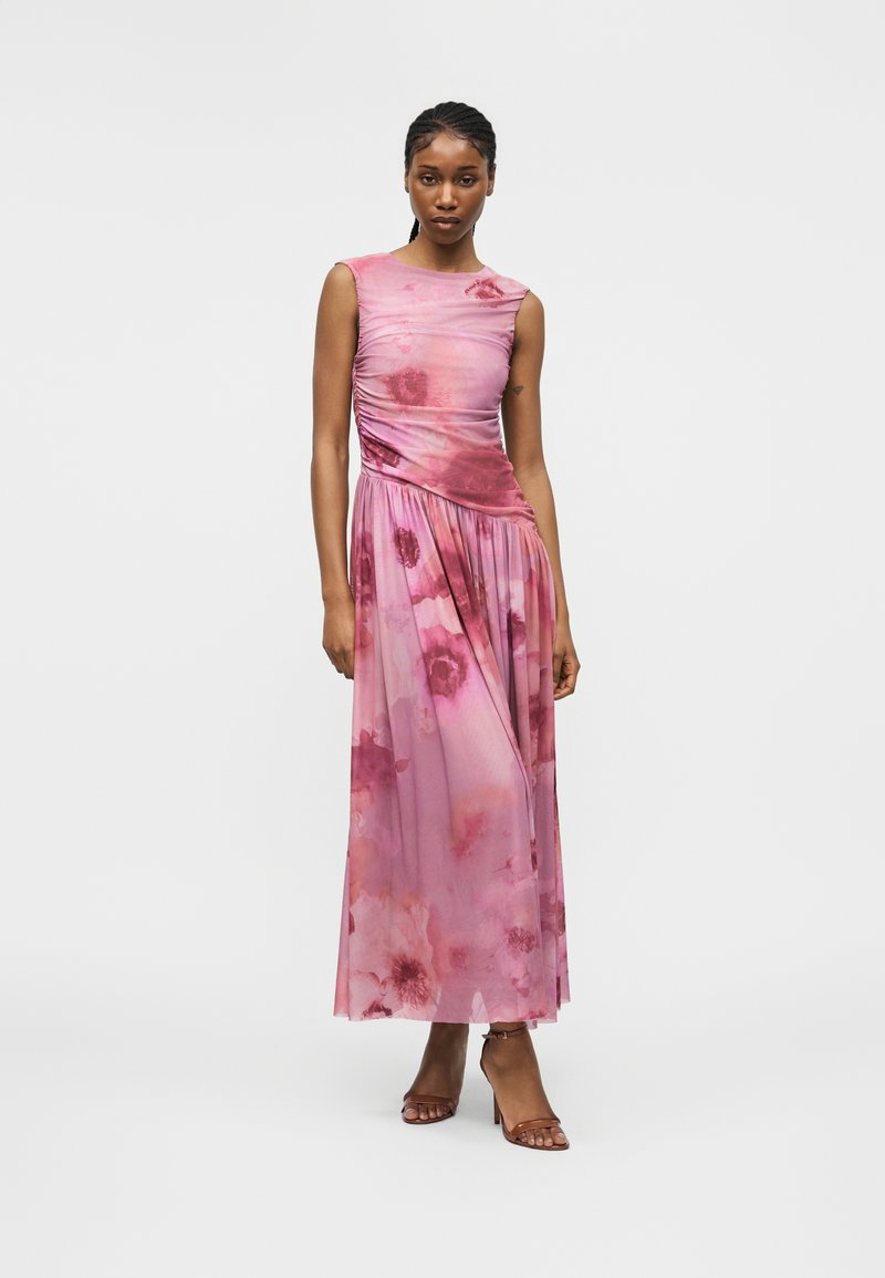 Femme debout portant une robe maxi rose à fleurs sans manches avec un corsage froncé et des sandales marron à talons hauts, sur un fond clair uni.