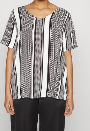 Person iført en sort og hvid, vertikalt striped kortærmet top med geometriske mønstre og sorte bukser, stående mod en ensfarvet baggrund.