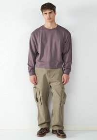 Langärmeliges, mauvefarbendes Baumwollhemd kombiniert mit khakifarbenen Cargo-Hosen. Die Hosen verfügen über große Seitentaschen und einen bequemen Schnitt. Braune Schnürschuhe sind ebenfalls enthalten.