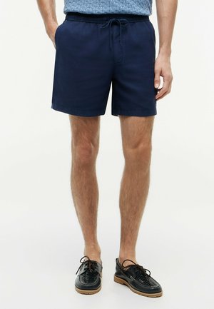 Man draagt marineblauwe shorts met trekkoord, een blauwe gestructureerde blouse en zwarte leren veterschoenen met rubberen zolen, staand tegen een effen achtergrond.