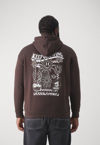 Uomo con felpa marrone con grafica di fiori, sole sorridente, api e testo "Continua così" sulla schiena, in piedi contro uno sfondo semplice.