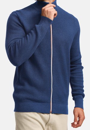 Mann trägt einen strukturierten blauen Pullover mit Reißverschluss, versehen mit weißen und roten Zippern, kombiniert mit beigen Hosen, vor schlichtem Hintergrund.