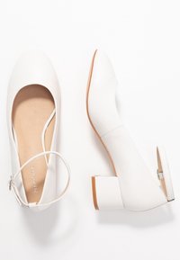 Vita pumpskor med rund tå, ankelrem och fyrkantig klack. Slät lädertextur med en beige sula och minimala detaljer.