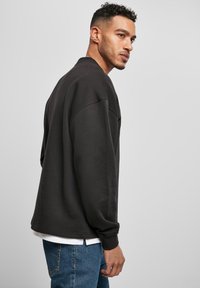 Hombre con cabello corto y rizado y barba, vistiendo una sudadera negra y jeans azules, mirando de lado contra un fondo claro y uniforme.