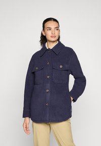 Kvinne i marinblå fleecejakke med knapped brystlommer og beige bukser, som ser litt til siden mot en enkel bakgrunn.