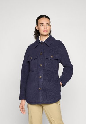 Vrouw in een marineblauwe fleecejas met knoopzakken op de borst en beige broek, die iets opzij kijkt tegen een effen achtergrond.