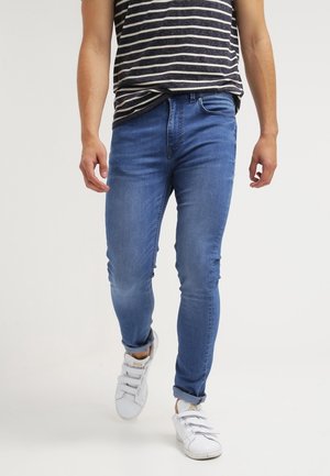 Homme portant un jean slim bleu, un t-shirt rayé noir et blanc, et des baskets blanches marchant sur un fond clair uni.