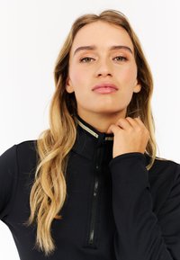 Sort langærmet top med høj krave og halv-zipped lukning, med en guldaccent omkring halsen. Glat, tætsiddende tekstur.