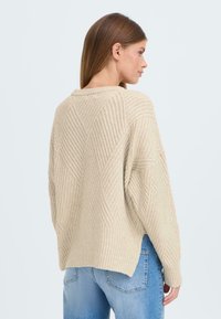 Beiger Pullover mit strukturiertem Rautenmuster und geripptem Saum. Bietet eine lockere Passform und überschnittene Schultern, kombiniert mit hellblauen Jeans.