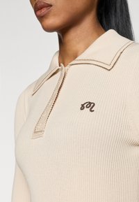 Beige ribbed knit top with a collar, featuring a zip closure and a brown embroidered logo on the chest. Smooth texture and tailored fit.