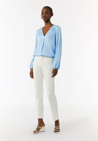 Himmelblause Langarmbluse mit V-Ausschnitt und gefältelten Details, kombiniert mit weißen Slim-Fit-Hosen und weißen Keilsandalen.