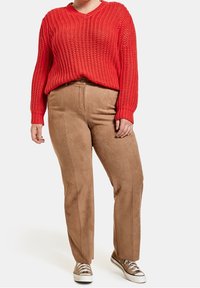 Osoba ubrana w czerwoną grubą sweterkę, jasnobeżowe spodnie z wysokim stanem i jasnobeżowe trampki stoi na białym tle.