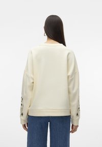 Sudadera de gran tamaño color crema con hombros caídos, con bordado floral en las mangas y puños acanalados. Combinada con pantalones de mezclilla.